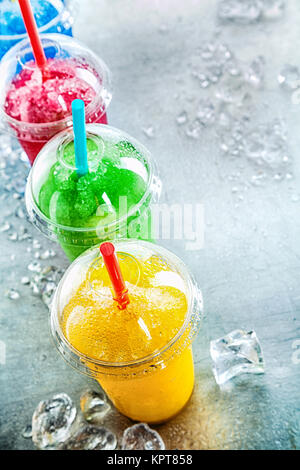 Hohen Winkel noch Leben Blick auf Bunte gefrorene Früchte Granita Slush Drinks in Kunststoff entfernt Becher mit Deckel und Trinkhalme angeordnet in der Zeile auf Kaltes Metall Oberfläche mit Eiswürfel und Kopie Raum Stockfoto