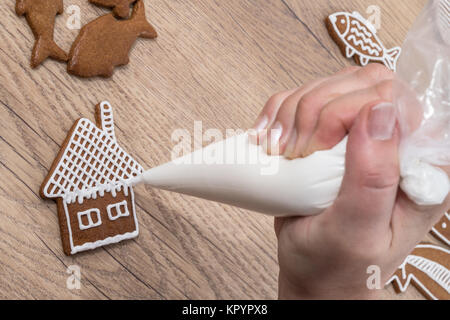 In der Nähe der weiblichen Hand, während die Dekoration Weihnachten Lebkuchen auf Holz. Die Frau ist die Malerei das Haus auf Keks mit Zuckerglasur Tasche mit Zucker Glasur Stockfoto