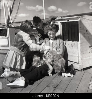 1950 s, historischen, "pet-friendly Travel" .... auf dem Deck eines Union-Castle Steamship Segeln für Südafrika, wir sehen Kinder sanft ein Hund von seiner Besitzerin statt Streicheln - die Pet scheint mehr in die Puppe interessiert - mit Stiften in - auf dem Deck als die drei Kinder. Stockfoto