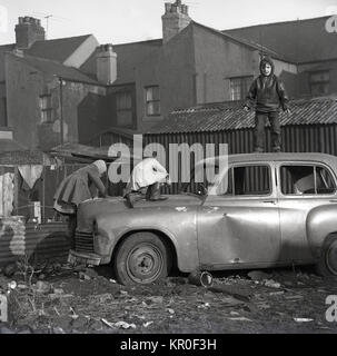 1960, städtische Spielplatz, schäbigen kleinen Kindern auf einem alten, verlassenen Auto auf den Inneren spielen - Stadt Ödland an der Rückseite der Reihenhäuser, England. Stockfoto