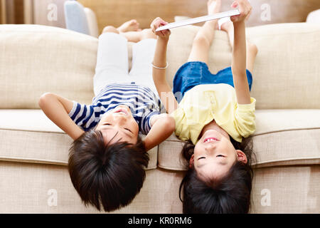 Zwei asiatische Kinder Jungen und Mädchen liegen auf den Kopf auf der Couch bei digital Tablet. Stockfoto