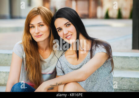 Zwei junge Schwestern sitzen auf der Treppe in der Straße und hören Musik. ein Mädchen mit langen brünetten Haar, eine Rothaarige, Mädchen, grau Shirt Stockfoto