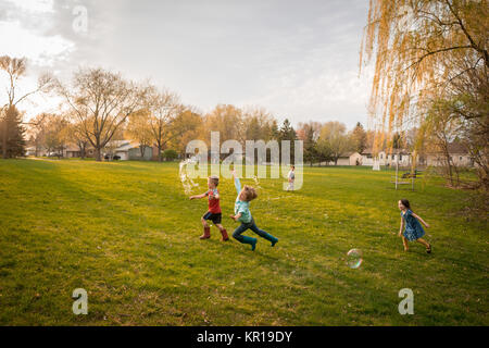 Vier Kinder jagen riesige Seifenblasen in einem öffentlichen Park Stockfoto