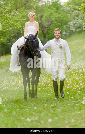 Hochzeit auf dem Lande. Die Braut, die in ihrem weißen Hochzeitskleid sitzt auf einem dunklen Pferd und wird durch Ihr Lachen Bräutigam geführt auf den blühenden Wiese. Stockfoto