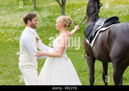 Hochzeit auf dem Lande. Die Braut, die in ihrem weißen Hochzeitskleid sitzt auf einem dunklen Pferd und ihr Bräutigam hilft Ihr abbauen, wenn das Pferd Stockfoto