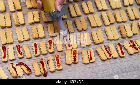 Die Süße Weihnachten Kekse mit Marmelade geklebt. Die Hand mit einem Metall Drücken und Schieben Marmelade auf das Gebäck auf Holz Hintergrund gelegt. Stockfoto