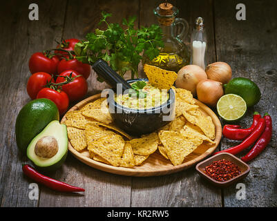 Schale mit leckeren hausgemachten Guacamole mit Nachos neben frischen Zutaten auf hölzernen Hintergrund Stockfoto