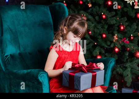 Süße kleine glückliche Mädchen mit Geschenk Box sitzen im Stuhl in der Nähe der Weihnachtsbaum. Horizontale Farbfotografie. Stockfoto