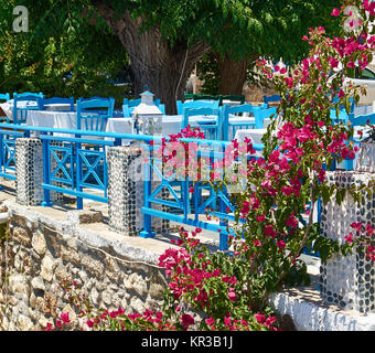 Terrasse im Sommer Restaurant in Griechenland, der Insel Rhodos. Blaue Stühle und Tische mit einem weißen Tuch unter einem großen Baum und das eisstockschiessen Bush mit hellem Lila Blüten an einer Steinmauer. Stockfoto