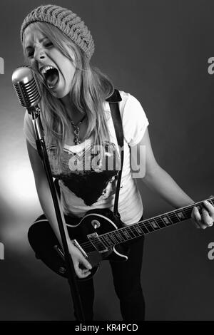 Schwarz-weiß Foto eines blonden Frau in ein altes Mikrofon schreien und spielen E-Gitarre. Hoher Kontrast mit Filmkorn hinzugefügt. Stockfoto