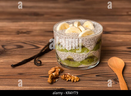 Chia Samen Pudding in Glas Schale mit Bananen, Kiwi, Walnüsse, Vanilleschoten und Löffel auf Holz- Hintergrund, raw food Konzept Stockfoto