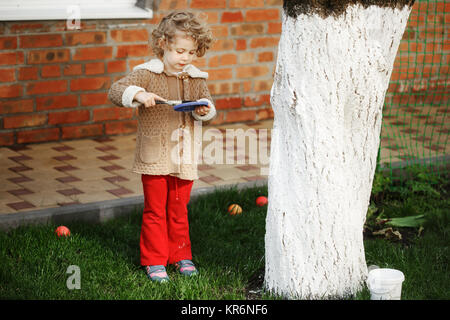 Wenig adorable blonde Mädchen, die den Baum mit weißer Farbe gegen Nagetiere im Garten im Frühling zu schützen. Stockfoto