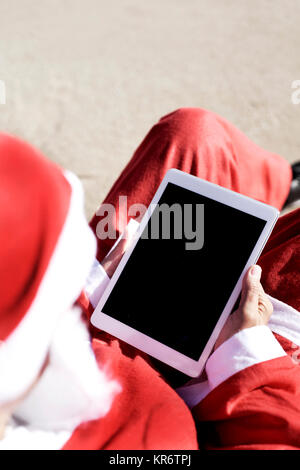 Nahaufnahme von Santa Claus mit seinem Tablet, mit einem schwarzen, leeren Raum in seinen Bildschirm, sitzen am Strand Stockfoto