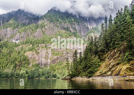 Sonne leuchtet auf einer Landspitze und einen steilen Berg Gesicht dahinter in Princess Louisa Inlet, während Wolken verdecken die hohen Gipfel beginnen sich zu heben. Stockfoto