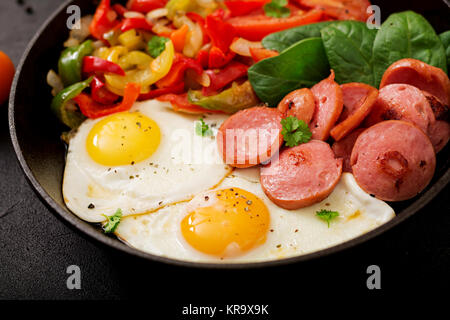 Frühstück. Spiegeleier mit Wurst und Gemüse in einer Pfanne auf einem schwarzen Hintergrund im rustikalen Stil. Stockfoto