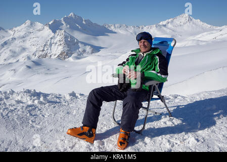 Mann im alpinen Skisport Kleidung sitzt in einer Chaiselongue und blickt auf die Berge Stockfoto