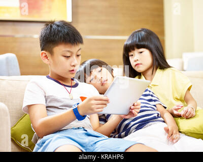 Drei asiatische Kinder zwei kleiner Junge und ein kleines Mädchen liegt faul auf der Couch spielen video game mit digitalen Tablet. Stockfoto