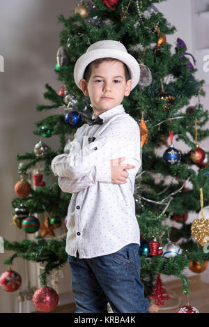 Junge mit ernsten Einstellung mit Kreuz die Arme in eine blaue Jeans und weißem Hemd mit schwarzer Schleife binden und festlichen weißen Hut feiern Weihnachten ständigen Alon Stockfoto