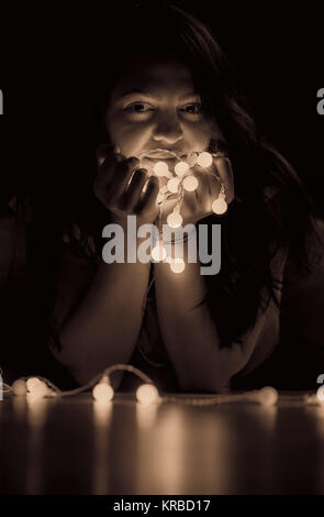 Sepia getont Portrait von eine junge attraktive Frau mit langem Haar sitzen auf dem Boden, in einem dunklen Zimmer, halten in ihren Händen ein String der Lichter. Stockfoto