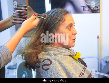 Stylist halten Sie einen Kamm bereit, eine reife Lady mit langen Haaren saß ein Salon Stuhl zu kämmen und mit einem Handtuch preid mit gelber Feder Stockfoto