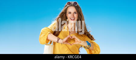 Perfekte Sommer. Lächeln gesunde Frau in kurzen Hosen und gelbe Bluse mit weißem Strand Beutel, der am Ufer des Meeres, die Herzförmigen Hände Stockfoto