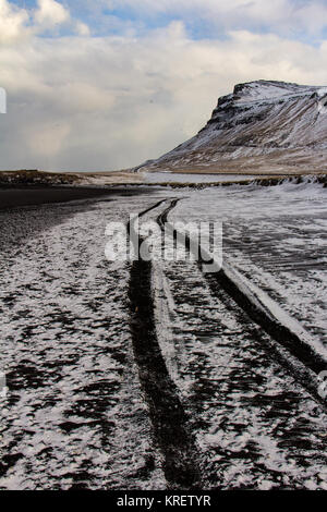 Schnee bedeckt schwarzer Sandstrand auf der Halbinsel Snaefellsnes in Island mit Reifenspuren in den Abstand auf einer Klippe führt. Stockfoto