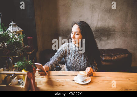 Schöne junge Mädchen verwendet, Typen text auf einem Mobiltelefon an einem Holztisch in der Nähe der Fenster und trinkt Kaffee in einem Cafe mit Weihnachten dekoriert. In einem grauen gestrickten Wollpullover angezogen Stockfoto