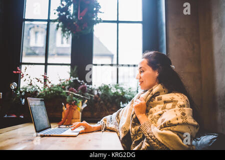 Schöne junge Mädchen nutzt laptop Technik, Typen text am Monitor in einem Café auf der Suche durch das Fenster an den hölzernen Tisch, im Winter mit Weihnachten dekoriert. in einem grauen gestrickten Wollpullover angezogen Stockfoto