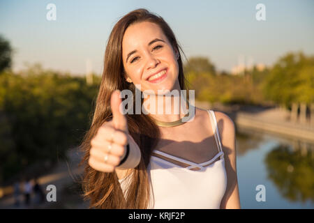 Glücklich aussehende junge Frau im Park an einem sonnigen Tag und deutete mit dem Daumen nach oben. Stockfoto