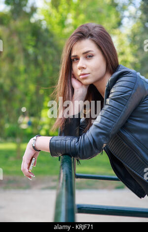 Junge Frau in schwarzem Leder Jacke holding Smartphone und lehnte sich auf Zaun an Kamera im Park. Stockfoto