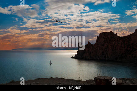 Abend Landschaft mit Sonnenuntergang und Wolken über einem ruhigen Meer, Klippen, Möwen und ein Segelboot, Krim, Novy Svet. Stockfoto