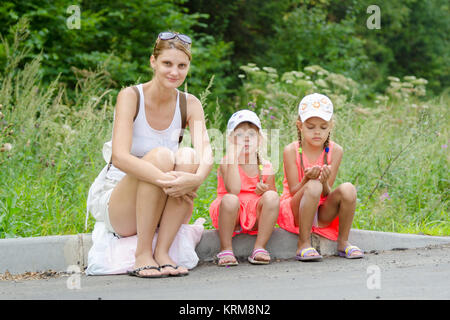 Mutter und zwei Kinder sitzen auf dem Bordstein der Straße wartete auf den bus Stockfoto