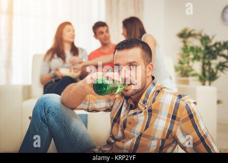 Junge lächelnde Mann Bier trinken zu Hause. Seine Freunde Spaß im Hintergrund. Selectiv konzentrieren. Fokus auf den Vordergrund. Stockfoto