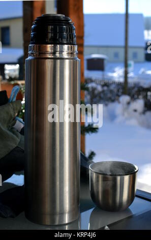 Eine Thermosflasche und einer Tasse Tee auf einem Glastisch und eine verschneite Szene im Hintergrund Stockfoto