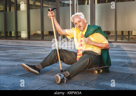 Rentner müde sitzen auf dem Skateboard, in der Hand einen Stock. Stockfoto