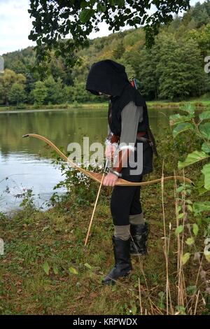 Mittelalterlicher Bogenschütze mit schwarzer Kapuze steht mit angespanntem Pfeil und Bogen auf dem See Stockfoto