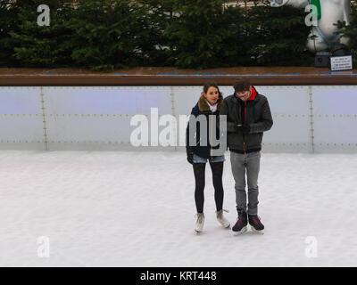 Junges Paar stehend auf Schlittschuhen Münchener EisZauber München Ice Magic Eisbahn Mann überprüfen Iphone für Nachrichten, die Freundin wartet geduldig entlang Stockfoto