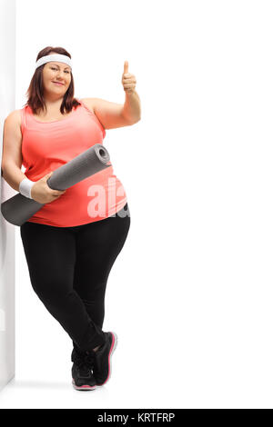 In voller Länge Porträt einer übergewichtigen Frau mit einem trainingsmatte einen Daumen oben unterzeichnen und lehnte sich gegen eine Wand auf weißem Hintergrund Stockfoto