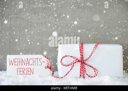 Ein Weihnachtsgeschenk auf Schnee. Zement Wand als Hintergrund mit Schneeflocken. Moderne und den urbanen Stil. Karte für den Geburtstag oder Seasons Greetings. Etikett mit deutschem Text bedeutet Weihnachtsferien Weihnachtsferien Stockfoto