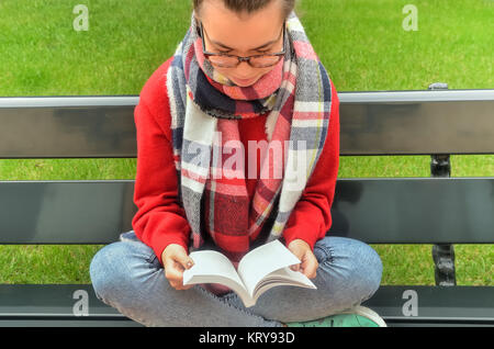 Asiatische Mädchen in der Brille sitzt auf einer Bank, kreuzen ihre Beine und liest ein Buch. Rund um den Rasen ist ein schönes grünes Gras. Ansicht von oben Stockfoto