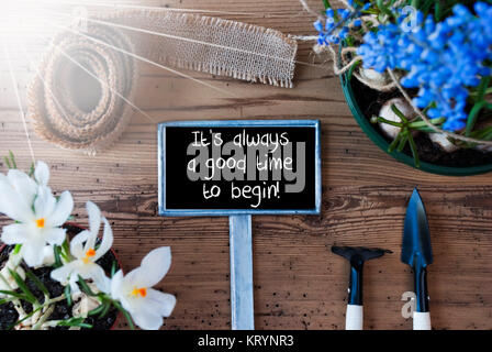 Mit englischen Zitat Zeichen Es ist immer ein guter Zeitpunkt, um zu beginnen. Sonnige Frühling Blumen wie Traubenhyazinthen und Krokus. Gartengeräte wie Rechen und Schaufel. Hanf Fabric Ribbon. Alter Holz- Hintergrund Stockfoto