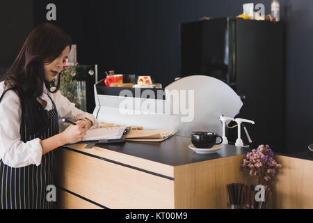 Inhaber eines kleinen Unternehmens mit Notebook an der Theke im Café. asiatische Frau barista Schürze tragen schreiben Hinweis an der Bar im Café. Food Service, Restaurant Stockfoto
