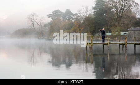 Ein Fotograf fängt Nebel, die aus dem ruhigen Wasser des Windermere See in Ambleside Pier, neben Herbst Wald in England Lake District Natio Stockfoto