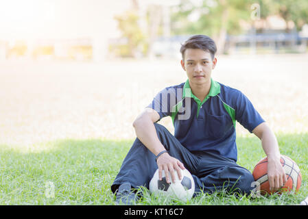 Smart Junge sitzt mit Fußball und Basketball auf grünem Gras, Sport und gesundes Konzept Stockfoto