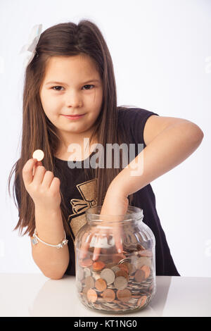 Eine sieben Jahre alte Mädchen, dass eine Cent Münze in einer Hand und mit der anderen packte Münzen in einem Glasbehälter. Stockfoto