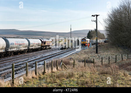 Zement Waggon Zug durch Class 66 Lokomotive geht ein anderer Zug laden Stein in das Abstellgleis im Bahnhof Ribblehead, North Yorkshire, Großbritannien gezeichnet Stockfoto