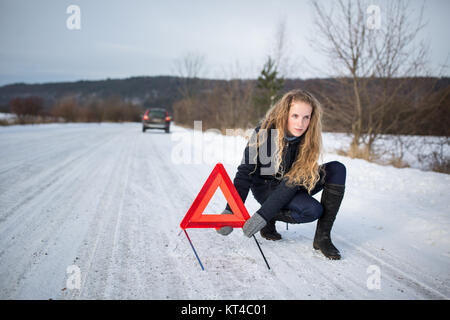 Junge Frau ein Warndreieck Einrichtung und Unterstützung gefordert, nachdem ihr Auto in der Mitte des nirgendwo an einem eiskalten Wintertag ist kaputt Stockfoto