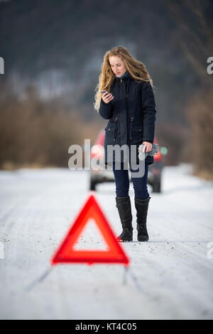 Junge Frau ein Warndreieck Einrichtung und Unterstützung gefordert, nachdem ihr Auto in der Mitte des nirgendwo an einem eiskalten Wintertag ist kaputt Stockfoto