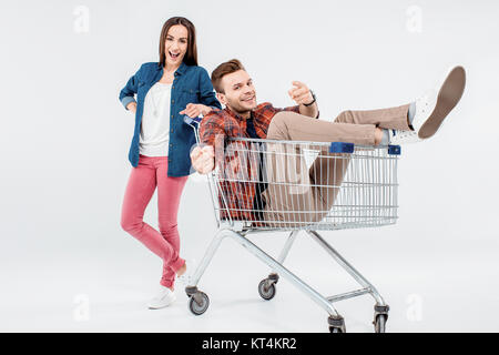 Lächelnde junge Frau stützte sich auf Einkaufswagen mit glücklicher junger Mann Stockfoto