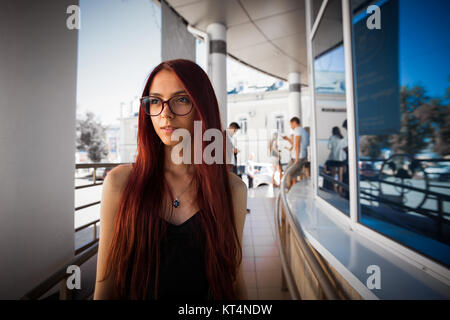 Stadt Portrait von Mädchen. Schöne Denken Mädchen mit langen roten Haaren und Brille beiseite. Hübsches Mädchen in schwarzem Kleid auf Stadt, schöne Architektur Hintergrund. Bild der hübschen Urban girl Stockfoto